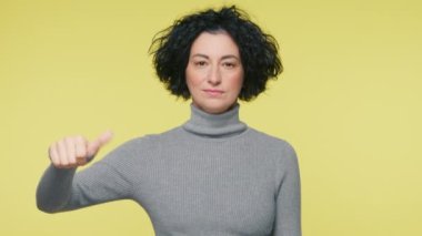 Smart woman 40s make choice, or makes decision thumb up or tnumb down, like or dislike, yes or no hand gesture slow mo. Mature woman in grey sweater making good-bad sign isolated on yellow background