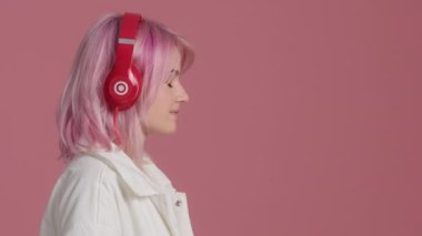 Serene young woman with pink hair with eyes closed wearing red headphones on pink background. Side view pretty student enjoys listening chill music audio sound meditating feeling no stress at studio