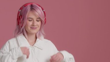 Young woman with pink hair wearing modern red headphones pointing the empty space for copy on pink background. Hipster student enjoys listening music audio sound meditating feeling no stress at studio