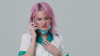 Young worried caucasian physician speaking on mobile phone with patient during workday in hospital. Slow motion studio background 4K doctor in lab coat protective glasses and stethoscope over her neck
