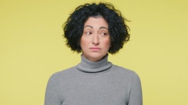 Middle age woman with thinking face expression standing over pale yellow background on studio wall. Slow motion portrait of confused woman, do not know what to do and how to react on weird situation