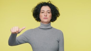 Pretty woman 40s makes choice decision thumb up or thumb down gesture, like or dislike, yes or no hand gesture slow mo. Mature woman in grey sweater making good-bad sign isolated on yellow background