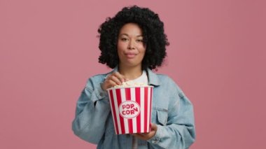 slow motion 4K Satisfied afro girl enjoying movie. Cheerful brunette African American woman of color in blue denim jacket eating popcorn while watching films isolated over pink background in studio