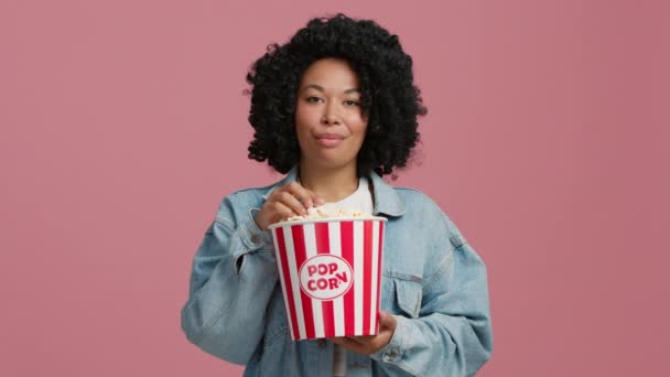 スローモーション 4K動画を楽しむアフロガールに満足 陽気なブルネットアフリカ系アメリカ人女性の色で青デニムジャケット食べるポップコーンながら見ますフィルム絶縁上ピンクの背景でスタジオ — ストック動画