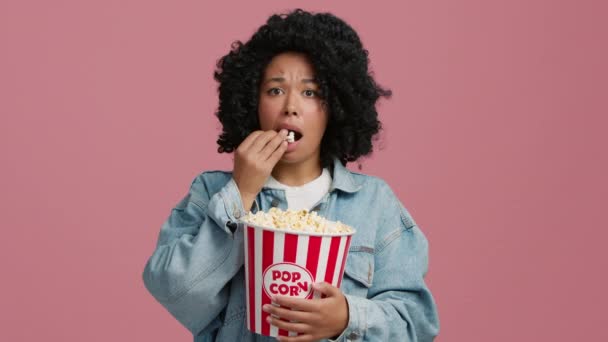 劇場で興味のある映画を見て魅力的な女性 スタジオで映画のコンセプトを見て巻き毛を持つアフリカ系アメリカ人女性 ピンクの背景にポップコーンを食べる集中モデル 週末時間 — ストック動画