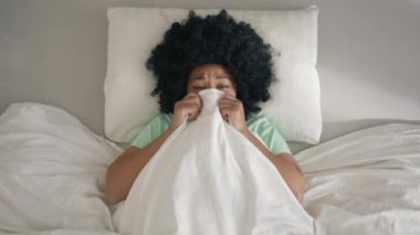 Frightened African American woman scared of ghosts on bed hiding under blanket at early morning. Top view on woman of color lying in bed and covering face by blanket to not see scary things at home 4K