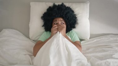 Adult african american woman scared by noise in home or hotel room in morning. Girl with afro hair infantility hiding under blanket, looking around scared to see monster or ghost slow motion nightmare