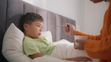 Sad little preschool boy at home with fever. Mom giving health care giving food to weak boy with no energy. African American mother caring for sick child lying in bed with flu, feeding him with spoon
