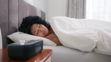 Happy woman of color sleeping in bed at home wakes up to digital alarm clock on bedside table to disable it. Girl stretches before getting out of bed to energize her body in morning in slow motion 4K