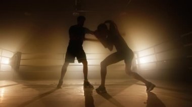 Close-up view of female athlete training in boxing club. Silhouettes of man and woman having intensive workout indoors. High quality 4k footage in golden orange yellow foggy light