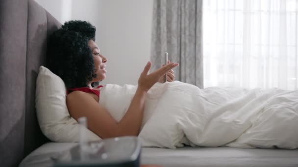 サイドビューの寝室のアパート 美しいアフリカ系アメリカ人の女性がベッドの中でスマートフォンを使用しています 家族の友達とビデオ通話で笑顔の女の子 スローモーション無線接続と通信 — ストック動画