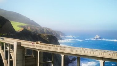 Slow motion following shot of bicyclist riding by world famous arch Bixby bridge, cinematic Pacific Ocean on motion background RED camera shot. Road trip scenic travel route. California vacation USA