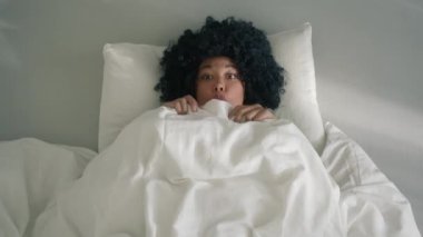 Goofy young african american girl making funny faces, being silly while lying in bed under blanket. Playful woman of color laughing and having fun in bedroom. Making kissy face and looking cross eyed