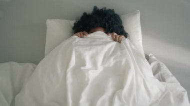 Portrait happy young woman of color peeks out from under blanket, smiles coquettishly, sticks out tongue, looks at camera. Girl black curly afro hair in bedroom. Relaxation, positive emotions concept