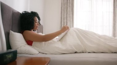Young woman of color reading book in cozy living room, turning page, learning, studying, education, read books, stay home concept. Beautiful African American woman awaken on sunny morning in bedroom