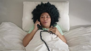 Afro girl spend time in bedroom. Top view of young relaxed woman of color in cotton pajama lying in bed with handset phone in hand actively discussing life, relationships, gossips or work with friend