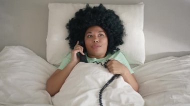 Indoor shot Portrait of dark haired curly woman in mint cotton pajama lying in kind size comfortable bed with handset phone talking with girlfriend discussing current gossips or relationships rumors