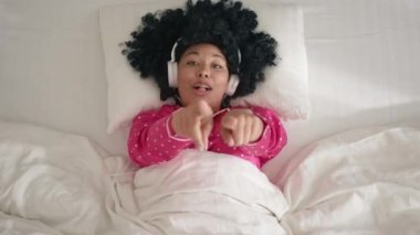 Joyful girl with afro hairstyle is dancing hands and singing wearing headphones having fun on bed at home. Modern entertainment, music and happy young people concept, slow motion funny woman of color