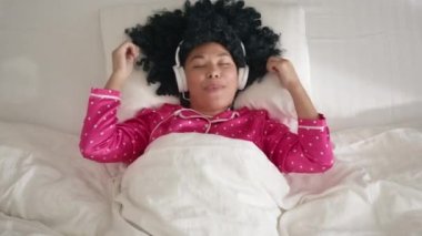 Modern entertainment, music and happy young people concept, slow motion funny woman of color. Joyful girl with afro hairstyle is dancing hands and singing wearing headphones having fun on bed at home