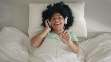 Excited happy young african mixed race woman wear wireless headphones having fun listen music in smartphone dance with hands lying on bed at home on sunny morning in slow motion