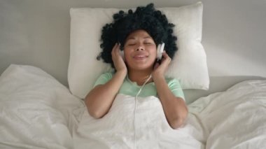 Young african american race woman relaxing on comfortable bed with wide smile wearing headphones. Woman of color enjoys listening chill music and audio sound relaxing at home. Top view in slow motion