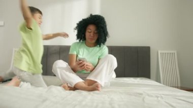 Happy african american mom and little son laughing, jumping on bed, looking at phone. Black mother using smartphone apps sitting on bed. Loving mommy holding cellphone playing with small kid at home