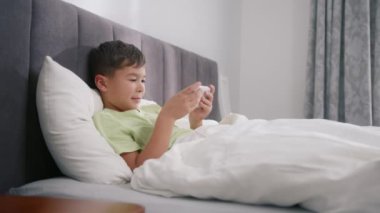 Preschooler playing mobile phone in slow motion. Kid using phone for gaming in bedroom. Child playing video game at home on sunny morning 4K. Mixed race boy playing mobile game on smartphone on bed