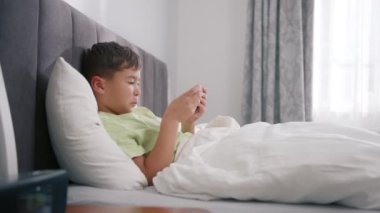 Kid using phone for gaming in bedroom. Child playing video game at home on sunny morning 4K. Mixed race boy playing mobile game on smartphone on bed. Preschooler playing mobile phone in slow motion