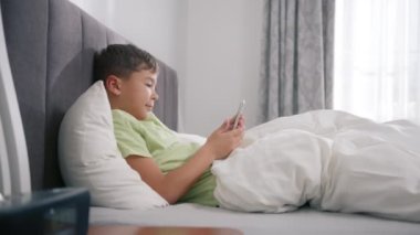 Happy mixed ethnicity little african american kid son holding phone having fun with smartphone. Child boy looking at cellphone, waving to camera on video call, enjoy using mobile apps at home in bed