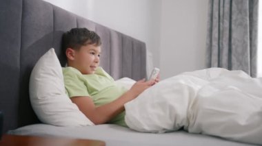 Child boy looking at cellphone, waving to camera on video call, enjoy using mobile apps at home in bed. Happy mixed ethnicity little african american kid son holding phone having fun with smartphone