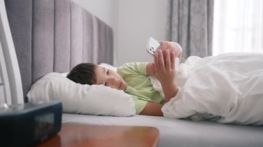 Cute mixed race kid boy holding smart phone enjoying using mobile apps, playing games at bed. Small mixed race child learning in cellphone, watching video, having fun with mobile technology concept 4K
