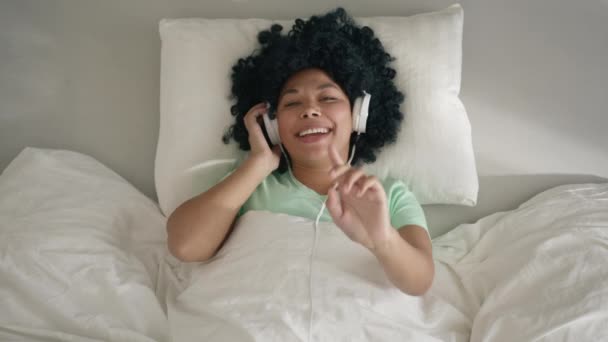興奮した幸せな若いアフリカの混合レースの女性は 遅い動きで晴れた朝に自宅でベッドの上に横たわっている手でスマートフォンダンスで楽しい音楽を聞いている無線ヘッドフォンを身に着けています — ストック動画