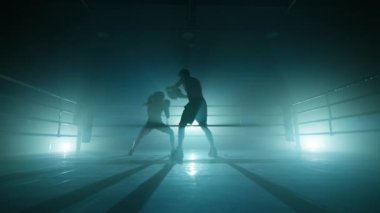 Unrecognized people punching in gloves in boxing ring. Man training female boxer to improve her body strength and endurance. High quality 4k footage in cold teal blue foggy back light