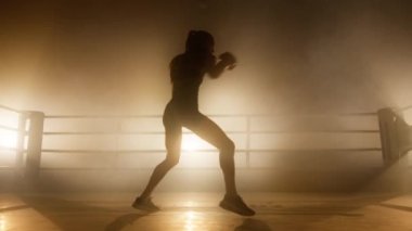 Caucasian sportswoman training in the boxing ring alone. Silhouette of a confident, strong girl working out in gloves at gym studio. High quality 4k footage in golden orange yellow foggy light