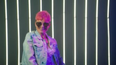 Active urban caucasian woman with short hot pink hair color in denim jacket dancing by neon lights background in disco club. Cool talented female dancer feeling free moves rhythmically hip-hop music