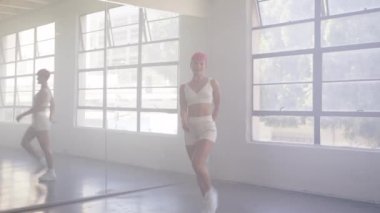 Smiling cheerful funky young energetic woman 20s years old in white tank top dancing and spinning in mirror studio background. Professional dancer walking to camera and performing combo movements 4K