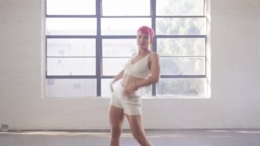 Female dancer performing dance show making freestyle stunts. Sexy stylish caucasian hipster girl. Fashion woman with hot pink hair and white outfit dancing inside white dance studio looking at camera