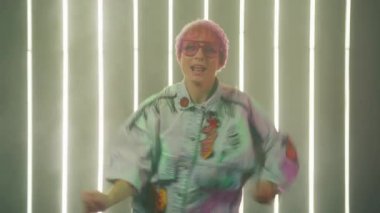 Happy young woman blogger performing dancer moves to music for sharing video on social media post stories at LED light wall background. Charismatic smiling positive girl in beanie funny trendy dance