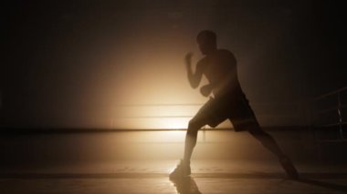 Silhouette of a kickboxer having an intensive workout in the gym. Unrecognized, strong man training in boxing club alone. High quality 4k footage in golden orange bronze foggy back light