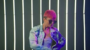 Closeup cheerful girl with bright magenta hair and modern sunglasses in fancy heart shape dancing with 80s disco moves at camera at nightclub. Beautiful woman clubbing at party neon lights background