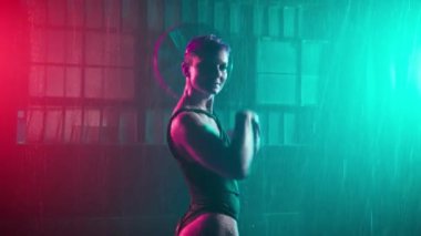 Stylish young girl dancing in grunge urban space, moving inside abandoned garage building under falling rain. Colorful studio spotlights. Half naked dancer on wet floor in futuristic cyberpunk space