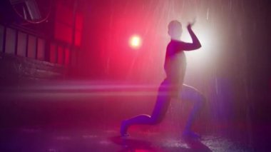 Passionate sexy professional dancer performing modern sensual dance in rain production studio environment with underground garage space and strobing neon red blue spotlights on motion background 4K
