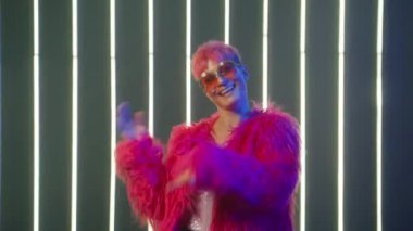 Funny smiling woman with pink hair having fun, smiling, dancing in studio against neon strip lights background. Funny girl in bright hot pink fur coat and sequin corset dancing by music in slow motion