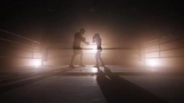 Silhouette of experienced coach working out with female boxer. Unrecognized couple training for body fit and endurance indoors. High quality 4k footage in golden sepia brown foggy back light