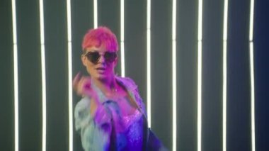 Girl moves to rhythm of music. Woman with colorful hairstyle dancing on LED lights background. Female in heart shaped sunglasses having fun, smiling beautifully to camera. Nightlife positive footage