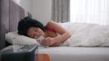 Beautiful African American woman using smartphone in bed at morning. Smiling girl scrolling social media feed. Slow motion wireless connection and communication shot. Side view bedroom in apartment