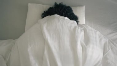 African American girl waking up in bed on early morning, feeling tired after sleepless night, lack of sleep and laziness. Woman of color looking at bright sun and closing eyes, hiding under blanket