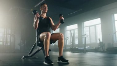 Close-up view of a man sitting while performing dumbbell curls. Portrait of active, strong sportsman targeting the biceps muscles on the front of his arms. High quality 4k footage