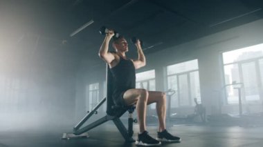 Close-up view of athlete holding dumbbells and lifting the weights above his head. Portrait of caucasian guy in his 30s training and targeting his arm and chest muscles. High quality 4k footage