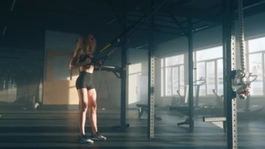 Portrait of female athlete developing muscular strength and endurance. Close-up view of caucasian woman squatting with a suspension trainer indoors. High quality 4k footage